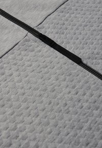 Tissu gris présentant un motif en losanges texturé, avec un détail de fermeture éclair noire le long du centre et un tirette de marque.