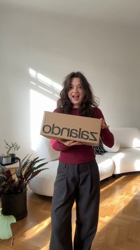 Kartónová krabica s čiernym logom "zalando", držaná osobou v burgundskej košeli a šedých nohaviciach, stojacou v dobre osvietennej miestnosti.