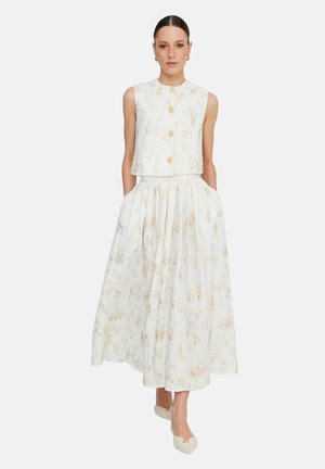 Frau trägt ärmelloses weißes Oberteil und passenden plissierten Rock mit hoher Taille und goldenem Blumenmuster, Hände in den Taschen und weiße Absätze mit goldenen Details.