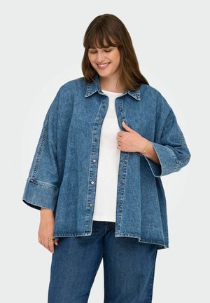 Modrá denimová košile s volným střihem, velké knoflíky, límec a široké rukávy. Nositá přes bílý top, v kombinaci s modrými džíny.