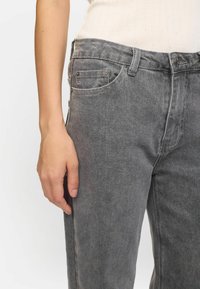 Graue Jeans aus Denim mit geradem Bein, fünf Taschen und sichtbaren Nähdetails; die Textur wirkt weich mit einem leichten Glanz.