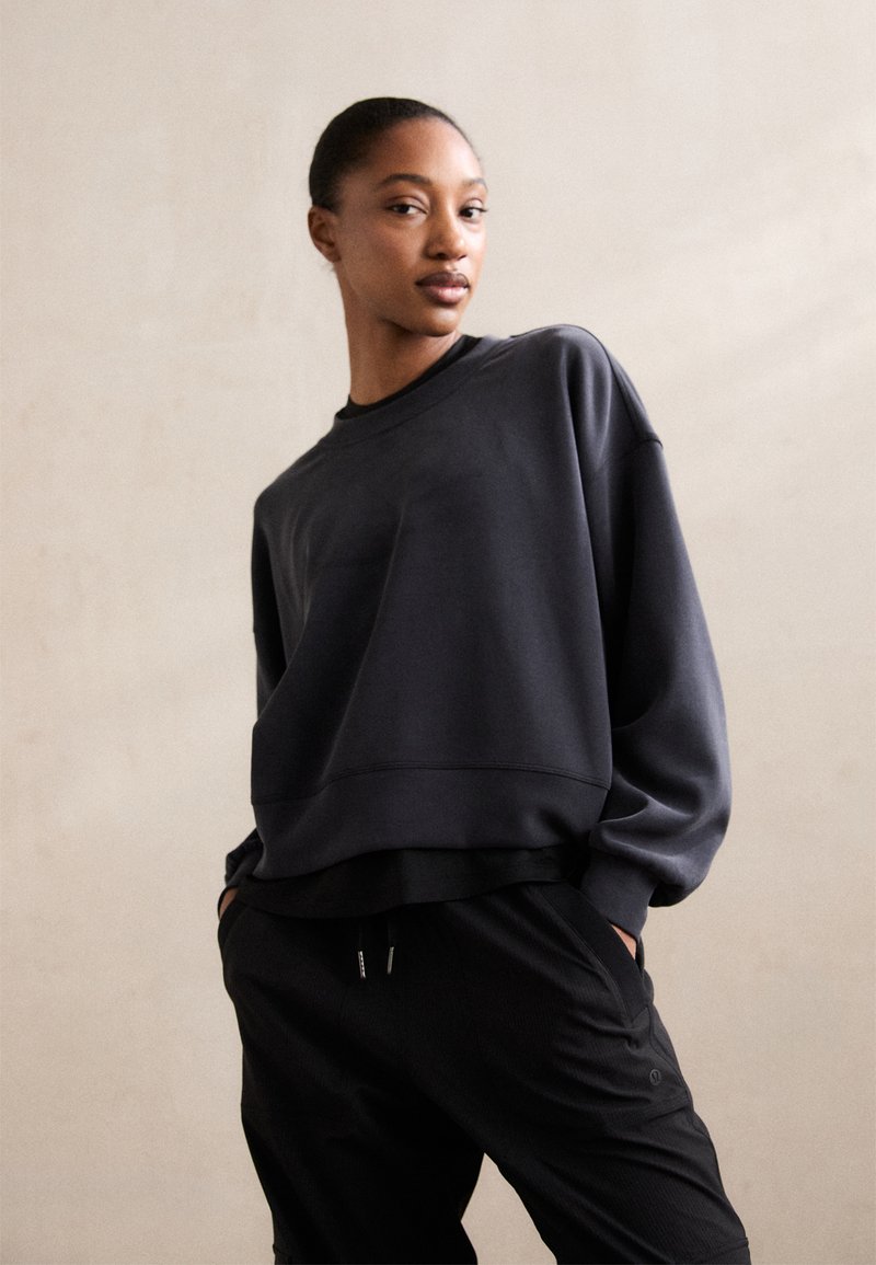 Fekete laza fitű pulcsi, kerek nyakkivágással és bordázott mandzsettával, fekete nadrággal párosítva. Simább anyag, látható minták vagy díszítések nélkül.