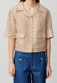 Kortärmad blus i beige tyg med röd blommönster, med krage och två framfickor. Kombinerad med blå denimbyxor.