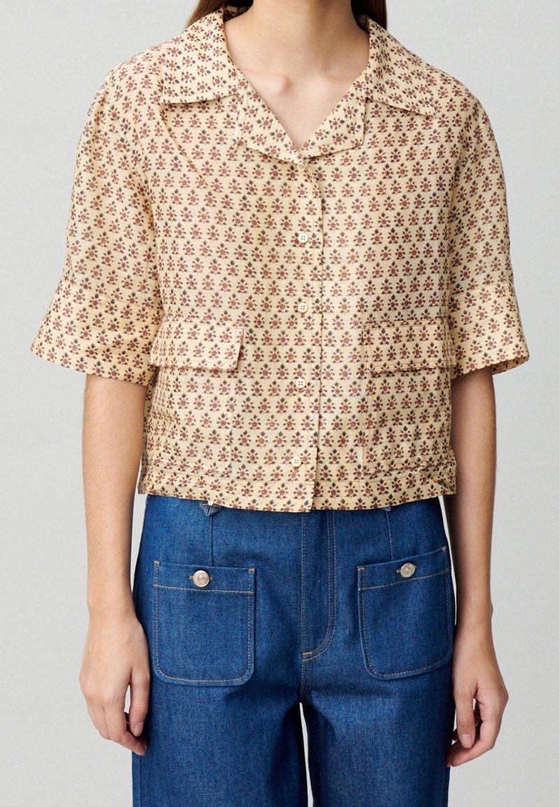 Kortärmad blus i beige tyg med röd blommönster, med krage och två framfickor. Kombinerad med blå denimbyxor.