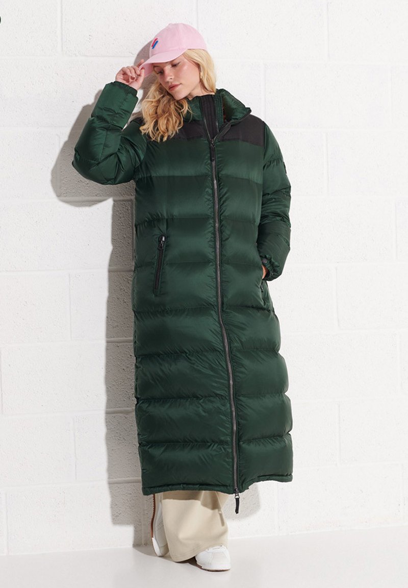 Zielony, pikowany długi płaszcz z zamkiem z przodu, czarną górną częścią, bocznymi kieszeniami, ściągaczami na rękawach oraz gładką teksturą. Noszony z różową czapką i białymi butami.