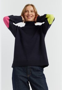Pull à col roulé marine en tricot, doté de coudières rose et verte, d'un ourlet côtelé et d'une coupe décontractée. Porté avec un denim foncé.