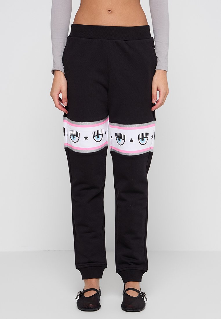 Schwarze Jogginghose mit einem horizontalen Streifen in Pink, Weiß und Silber, verziert mit Augen- und Sternmustern. Mit Bündchen an den Knöcheln.
