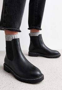 Czarne skórzane botki Chelsea z elastycznymi panelami bocznymi, płaskimi obcasami i białymi przeszyciami. Noszone z szarymi prążkowanymi skarpetkami i czarnymi skróconymi dżinsami.