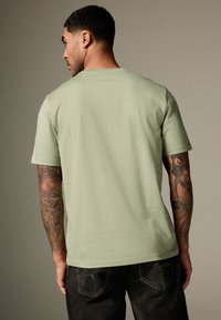 Svetlo zelena majica s kratkimi rokavi, sproščenega kroja, mehke teksture in klasičnega okroglega izreza. Predstavlja tetovirane roke in temne jeans hlače.