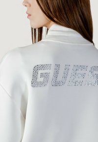 Sudadera blanca con un gran logo "GUESS" en la espalda hecho de pedrería plateada, con una textura suave y un ajuste relajado.