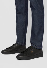 Sneakers in pelle nera con suole in gomma flat, punta arrotondata e design con lacci. Presentano un branding sottile arancione. Indossati con jeans blu scuro.