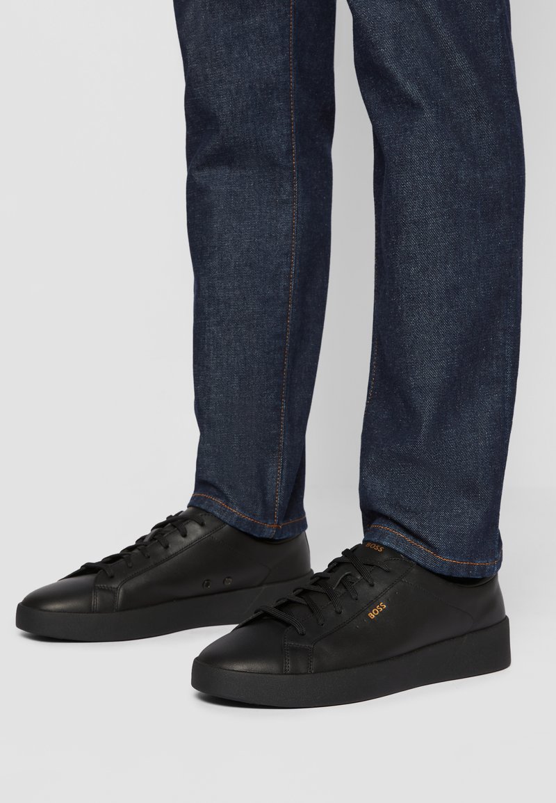 Sneakers in pelle nera con suole in gomma flat, punta arrotondata e design con lacci. Presentano un branding sottile arancione. Indossati con jeans blu scuro.