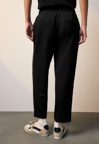 Juodos siaurėjančios kelnės su lygiu paviršiumi, turinčios dvi galines kišenes. Dėvimos su smėlio spalvos sportbačiais, pabrėžiantys aptakų, kasdienį dizainą.