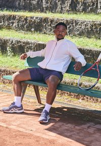 Hvid lynlåslukket jakke, marineblå shorts, grå tennissko, siddende på en grøn bænk ved siden af et rødt tennisketsjer, på en tonesandbane med sten trin.