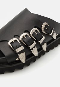 Sandale glissante en cuir noir avec quatre accents de boucles en métal argenté, semelle crantée et design supérieur lisse. Les détails matériels texturés sont prédominants.