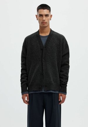 Homme aux cheveux courts et foncés portant un cardigan foncé boutonné, une chemise bleue et un pantalon large foncé, debout devant un fond clair uni.