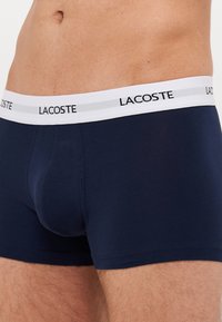 Tamno plavi pamučni bokserice s bijelim pojasom označenim "LACOSTE." Imaju uski kroj i glatku teksturu, bez vidljivih uzoraka ili akcenata.