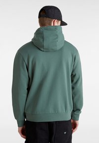 Felpa verde con cappuccio, tasca a marsupio, polsini e orlo a costine. La texture sembra morbida; presenta una vestibilità rilassata. Indossata con un berretto nero.