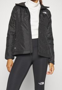 Svart isolerad jacka med huva, quiltat mönster, dragkedjestängning och The North Face-logotyp på vänster bröst, i kombination med svarta leggings.