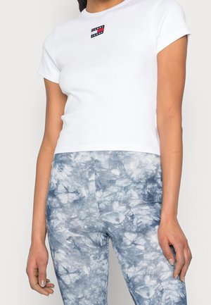 Wit T-shirt met korte mouwen en een klein rood, wit en blauw logo op de borst, gecombineerd met blauw tie-dye leggings met verticale strepen.