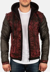 Giacca di jeans rossa e nera con zip frontale, maniche in pelle, dettagli con borchie e cappuccio. Presenta una texture consumata e accenti metallici.