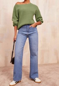 Pull vert en maille à design épaules dénudées, associé à un jean large bleu clair et des baskets blanches à rayures noires.