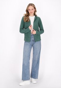 Hoodie in pile verde con zip completa, abbinato a jeans azzurri chiari e sneakers bianche. Texture morbida, vestibilità casual e design con cappuccio.