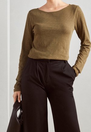 Person trägt ein olivgrünes Langarmoberteil und schwarze, weit geschnittene High-Waist-Hosen und hält eine schwarze Handtasche in einer Hand.