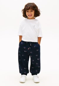 Χαμογελαστό παιδί στέκεται με τα χέρια στις τσέπες ενός σκούρου μπλε παντελονιού με σχέδιο μπάσκετ, φορώντας λευκό t-shirt και λευκά αθλητικά παπούτσια σε λευκό φόντο.