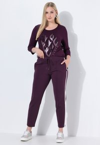Jonge vrouw in een paarse top met vlammenprint en bijpassende broek met een witte streep, staand in een casual houding tegen een witte achtergrond.
