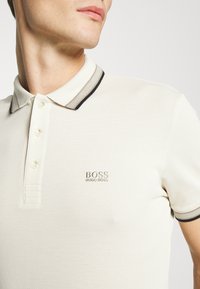 Krämfärgad pikétröja i texturerat tyg med svart- och grårandig krage, tre knappar och en broderad "BOSS"-logotyp.