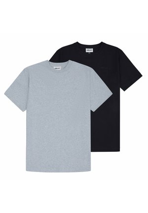 Jonge man met krullend haar die een effen lichtgrijs T-shirt met korte mouwen en ronde hals draagt en een donkere broek, staand met de handen achter zijn rug.