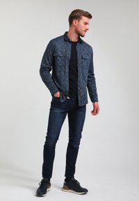 Majica s dolgimi rokavi v mornarskem vzorcu z dvema žepoma na prsih, v kombinaciji s temnimi jeans hlačami in temnimi teniskami, postavljena na enostavni beli ozadju.