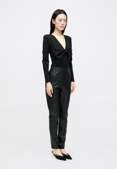 Μαύρη μπλούζα με μακριά μανίκια και βαθύ V-λαιμόκοψη, διακοσμημένη με φιόγκο, συνδυασμένη με εφαρμοστά μαύρα δερμάτινα παντελόνια και μυτερές μαύρες μπαλαρίνες. Λείο υφή σε όλη την επιφάνεια.
