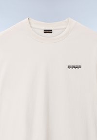 Lys beige bomulds t-shirt med rund hals, korte ærmer og et lille sort logo på venstre bryst. Blød tekstur, enkel design.
