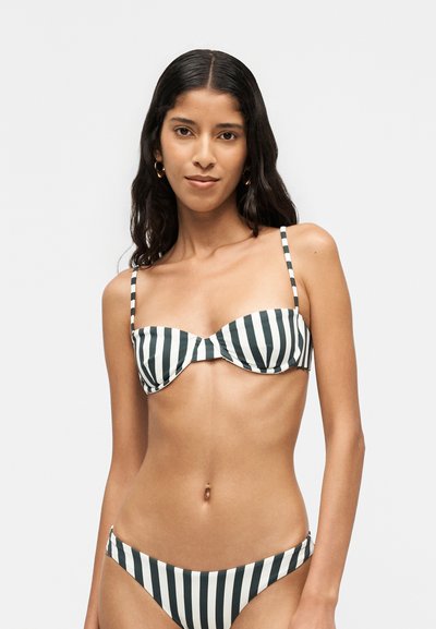 Femme aux longs cheveux foncés portant un haut et un bas de bikini à rayures verticales noires et blanches, debout devant un fond clair uni.