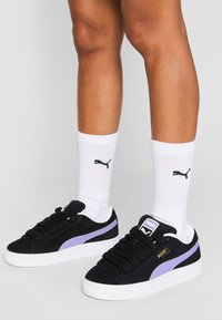 Baskets en daim noir avec des accents violets, semelle blanche et lacets noirs, associées à des chaussettes blanches arborant un logo Puma noir.