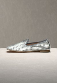 Ασημί μοκασίνι από διάτρητο δέρμα με στρογγυλεμένη μυτερή μύτη και χαμηλό τακούνι, σε ουδέτερο φόντο και επιφάνεια.