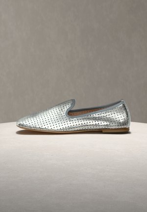 Sølvfarget perforert lær slip-on lavsko med avrundet spiss tå og lav hæl på en nøytral overflate og bakgrunn.