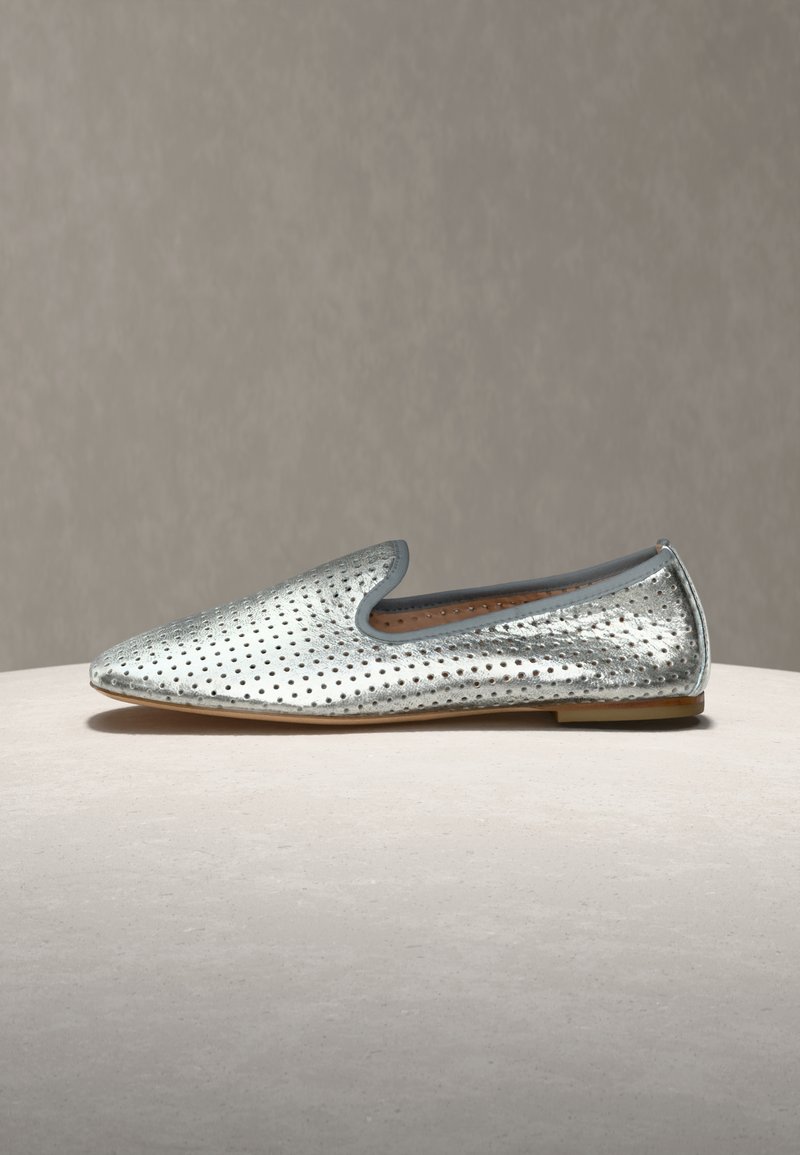 Chaussure plate en cuir perforé argenté, à enfiler, avec un bout arrondi pointu et un petit talon, posée sur une surface et un fond neutres.