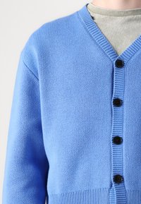 Cardigan albastru deschis realizat dintr-un material tricotat, având decolteu în V, butoane negre și tiv cu nervuri. Textură de cusătură vizibilă.