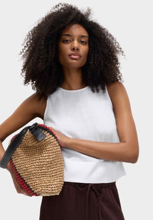 Sac en paille tissée de forme courbée, accents rouges et noirs, avec une poignée en cuir noir. Haut sans manches blanc présenté avec un pantalon marron.