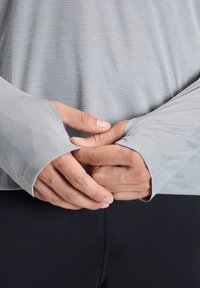 Light gray long-sleeve shirt with a ribbed texture and thumbholes, paired with black pants. Focus on hands positioned at the waist.