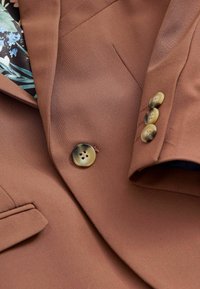 Blazer marron avec quatre boutons en écaille de tortue sur la manche, un bouton à l'avant, et une doublure à motifs floraux visible au niveau du col.