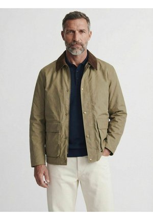 Mand med gråt skæg iført beige jakke med brun krave, navy skjorte og hvide bukser, stående mod en ensfarvet lys baggrund.