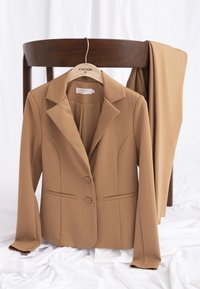 Blazer beige cintré sur un cintre en bois et pantalon assorti posé sur une chaise en bois foncé sur un fond en tissu blanc.