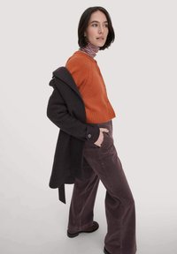 Oranger Strickcardigan, der über einem bordeauxroten Mantel getragen wird, kombiniert mit weiten Cordhosen. Ein gestreifter Rollkragenpullover ist am Hals sichtbar.