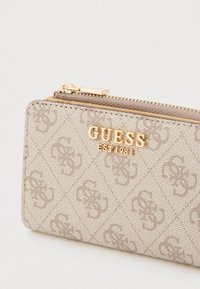 Bēšas krāsas maka ar rāvējslēdzēju, kurā ir iegravēts logotipa raksts, zelta krāsas metāla detaļas un gluda tekstūra. Kompakts dizains ar sānu rāvējslēdzēja aizdari.