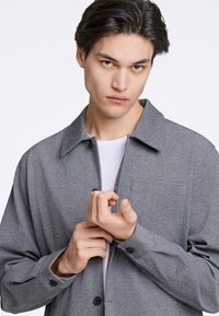 Graue knopfverschlossene Jacke mit Kragen, ausgestattet mit einer Tasche und strukturiertem Stoff. Über einem weißen Hemd getragen, Hände in der Nähe des Kragens platziert.