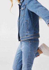 Ljust blår denimjacka med framfickor, knäppningar och matchande ljusblå jeans. Vita sneakers med texturerade sulor.